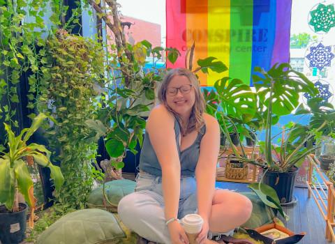 Abby sits cross-legged and smiles at the camera with eyes closed and coffee in hand in front of a colorful striped flag.